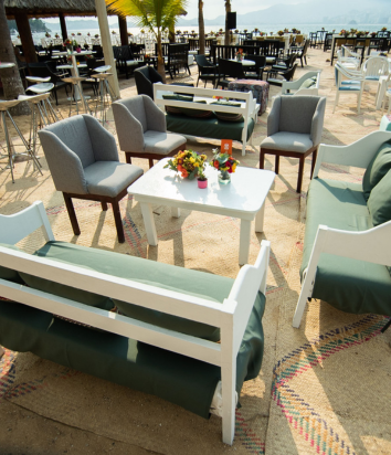Área lounge en la playa con muebles blancos y grises, decorada con cojines verdes y arreglos florales, ideal para eventos al aire libre.