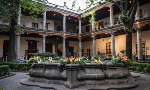 Patio colonial con una fuente central decorada con flores coloridas y velas, rodeado de jardines y arquitectura tradicional.